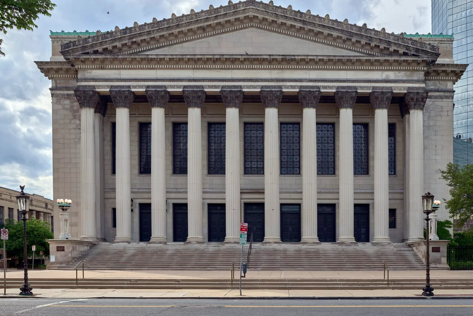 The Symphony Hall facade head-on from Court Street in Springfield on an overcast spring afternoon — a limestone-clad Classical-Revival front with six tall fluted columns rising to a deep dentilled pediment, three pairs of bronze-framed double-height windows set between the columns, a row of carved doorways at street level above a wide stone stair, ornate cast-iron lampposts flanking the entry, an empty sidewalk and curb in the foreground, a sliver of a modern glass tower at the right edge, and a soft pale-grey sky above.