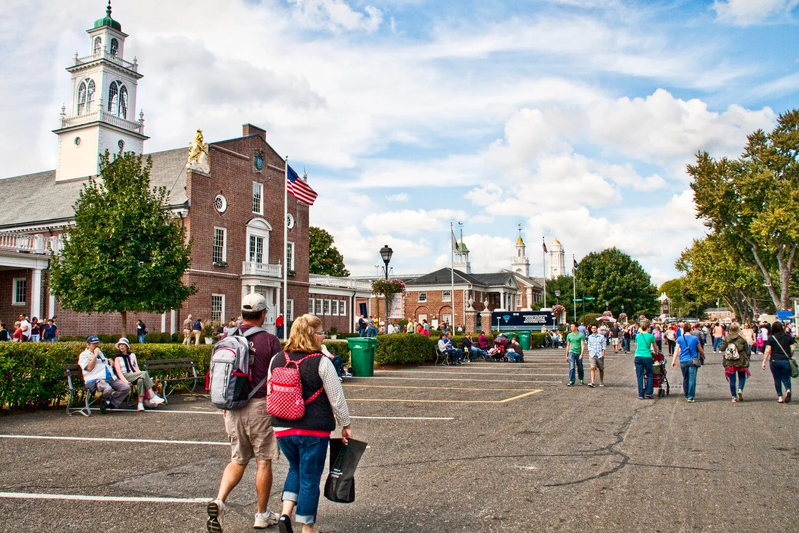 The Avenue of States at the Big E in West Springfield on a partly-cloudy late-summer afternoon. A red-brick Massachusetts state-house replica on the left with a tall white-columned cupola, a clock face, and a gilded sculpture above its pediment, an American flag on a pole at the entrance, fairgoers in shorts and backpacks walking down a wide paved avenue, smaller cupolas and white-trimmed gables of the Connecticut and Rhode Island replicas farther down the row, leafy green trees framing the right edge, and a bright blue sky scattered with cumulus clouds above.