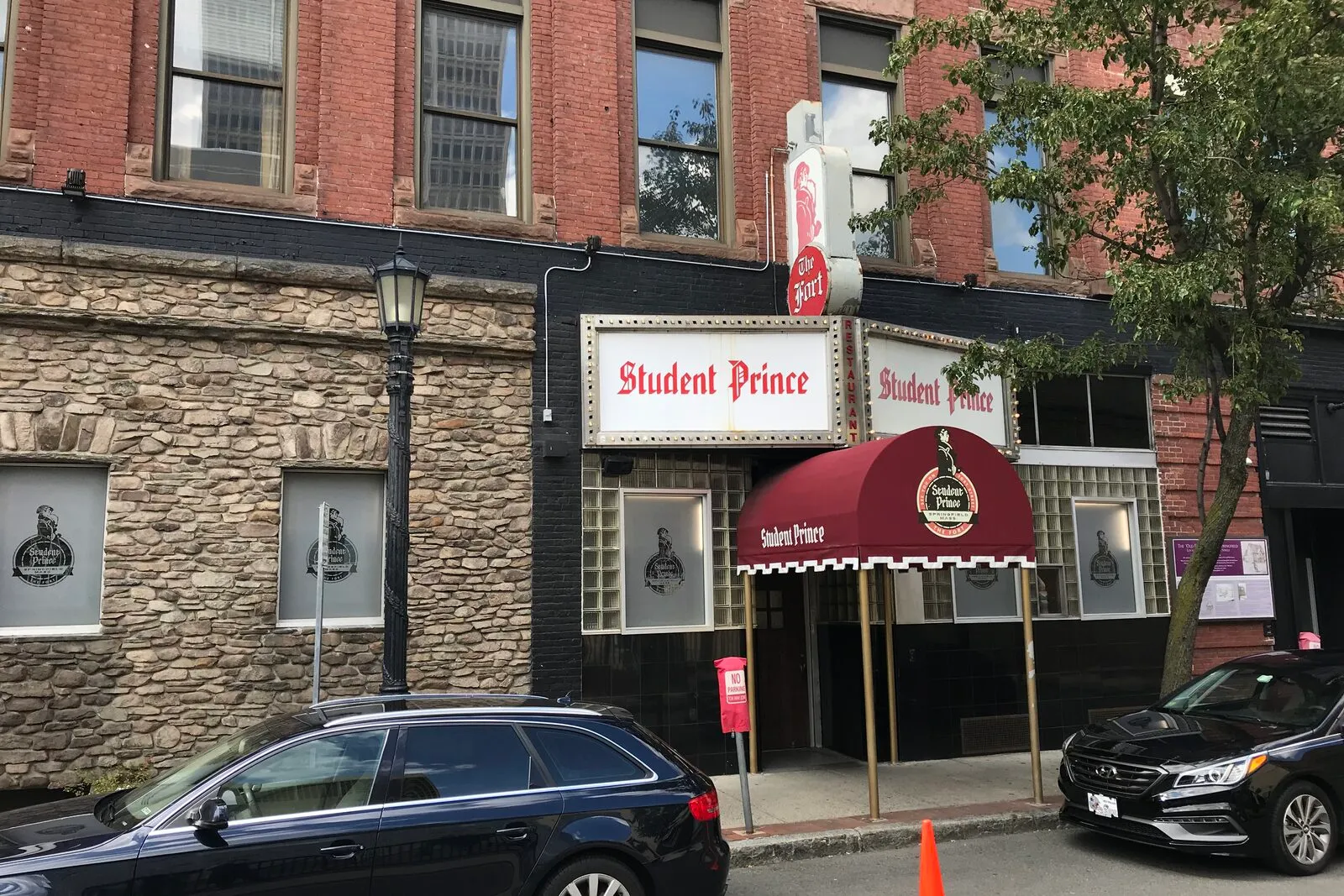 The Student Prince storefront on Fort Street in downtown Springfield on an overcast late-summer day. A two-story building with a rough fieldstone-faced ground floor and red-brick upper floors, two white sign panels reading 'Student Prince' in red blackletter lettering above the entry, a vertical neon blade sign with a beer-stein illustration mounted high on the brick, a red barrel-vault awning over the doorway carrying the Student Prince crest, flanking display windows holding silhouetted stein figures, a black wrought-iron lamppost and post-mounted lantern beside the entry, and parked cars and an orange traffic cone along the curb in the foreground.