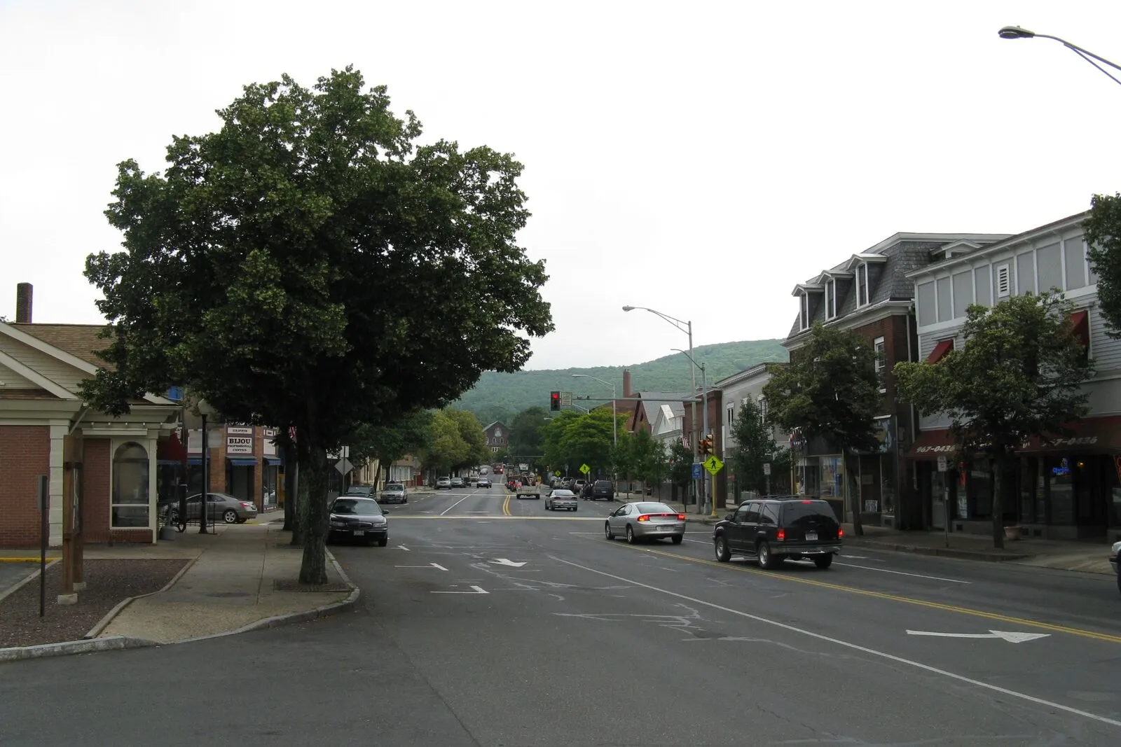 A summer view down Main Street in downtown Ware. A row of late-19th-century brick commercial blocks on the right with storefronts at street level and tall arched upper-story windows, a line of mature street trees in full green leaf along the curb, parked cars on both sides of the wide street, a few pedestrians on the sidewalk, and the road running off into the distance under a partly cloudy blue sky with a low ridge closing the view at the end.