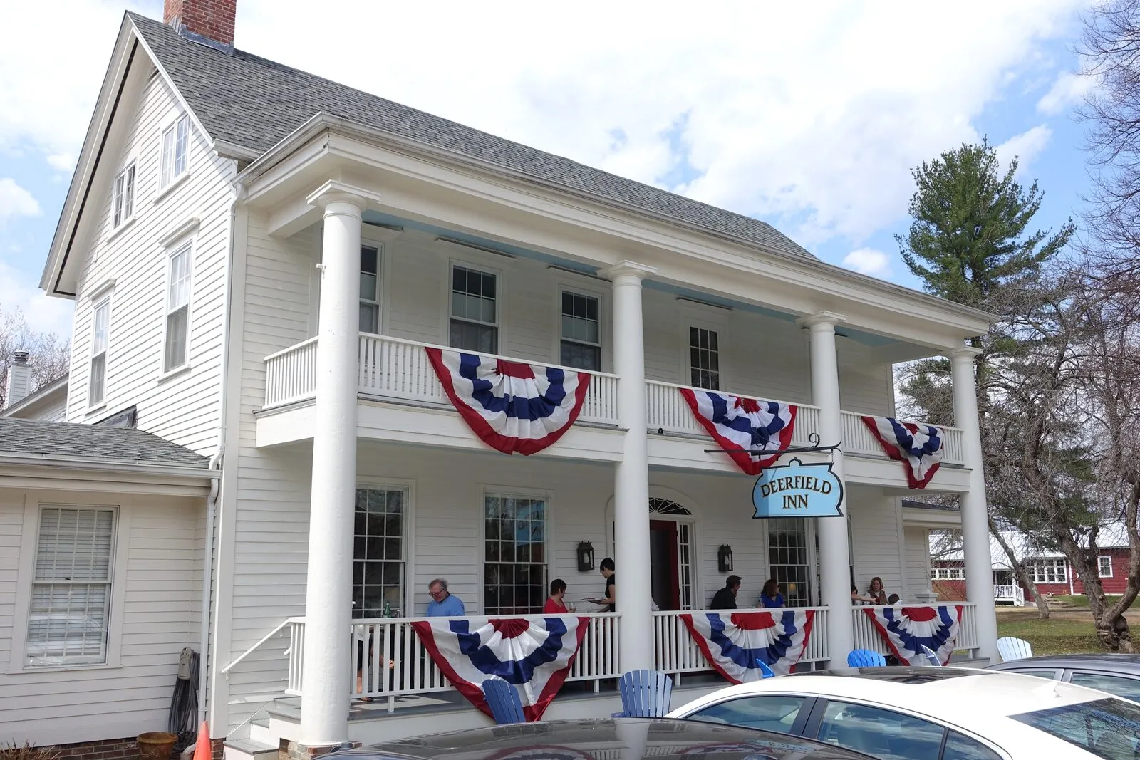 The Deerfield Inn on The Street in Old Deerfield — a two-story white clapboard building with a two-tier columned porch, patriotic red-white-and-blue bunting draped along the porch rails, a small hanging sign reading Deerfield Inn, and guests chatting on the porch in early-spring light.