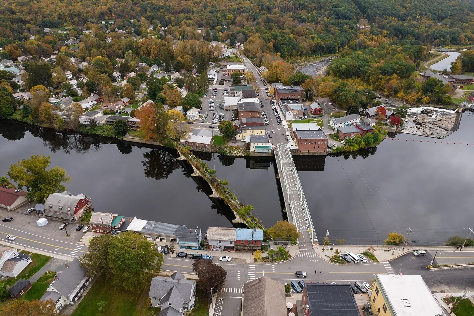An aerial view of the Deerfield River at Shelburne Falls in mid-October, looking south across the village — the river running across the foreground over the bedrock-and-cobble bed of the falls, the steel Iron Bridge carrying Bridge Street over it on the right, the planted Bridge of Flowers running parallel just upstream on the left, the West End Pub building and outdoor deck at the foot of the Iron Bridge on the near (Buckland) bank, and the dense hilltown forest of the Deerfield valley in late-fall color rising on both sides of the river.