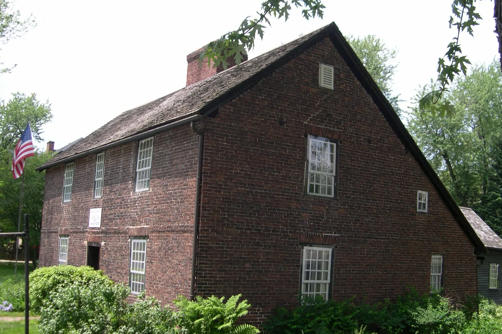 The Josiah Day House on Park Street in West Springfield on a sunny early-summer afternoon: a 1754 two-story brick saltbox house with a steeply pitched asymmetric roof sweeping low to the rear, deep-red weathered brick walls, a tall central chimney, six-over-six paned windows in white frames, a small entry door with a US flag on a pole at the corner, lush green ferns and shrubs banked along the foundation, a few mature deciduous trees framing the right edge with full summer foliage, and a partly cloudy sky overhead.
