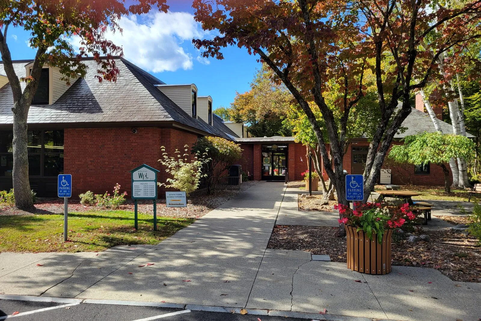 The front entrance of the Wilbraham Public Library on a sunny October afternoon: a low-slung brick building with a steeply pitched dark slate-grey roof, a recessed glass-fronted entry vestibule between two brick wings, two small accessibility-parking signs and a notice-board flanking the walk in the foreground, a black metal trash receptacle and a wooden picnic-table bench at right beside a small Japanese maple in full red-orange fall color, taller maples in mixed green-yellow-red foliage arching overhead, a striped asphalt parking lot in the immediate foreground, and a deep blue sky with a few thin white clouds visible between the leaves.