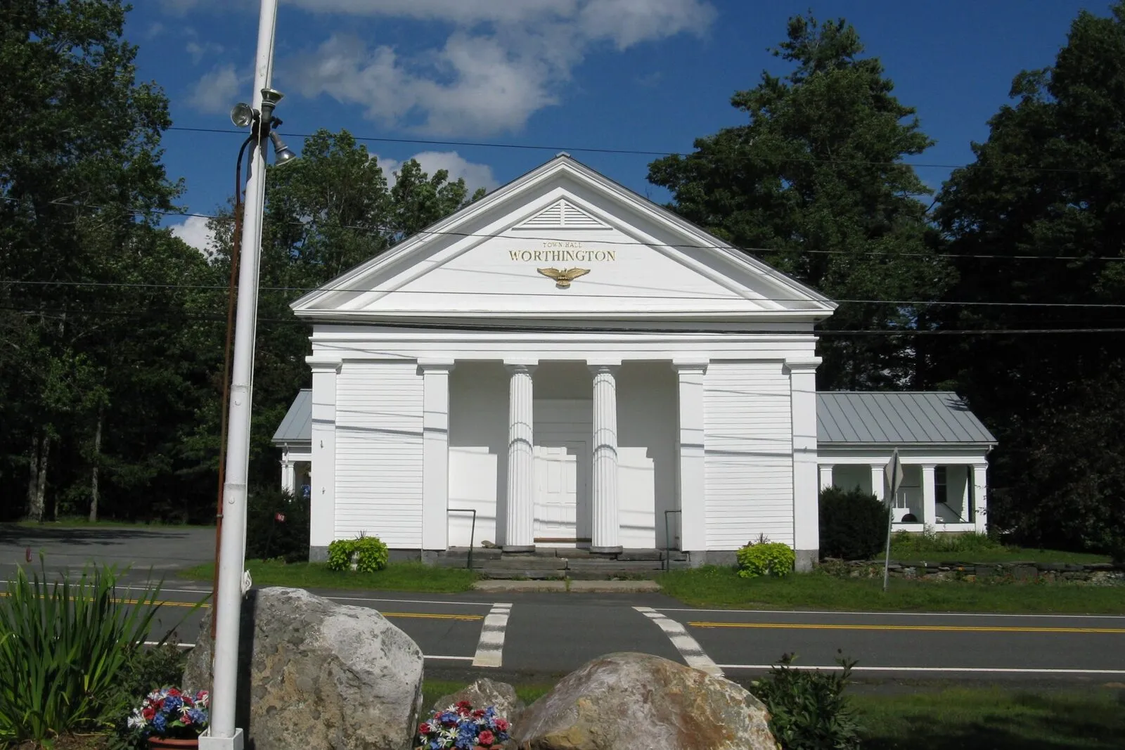 The Worthington Town Hall in Worthington Center on a sunny late-summer afternoon. A small white-clapboard Greek Revival civic building with a low-pitched gabled roof forming a full pediment across the front, four square Doric columns supporting the porch, three recessed entry doors set behind the columns, a gilded sign reading WORTHINGTON over the central entry, a metal standing-seam roof on the lower side wing to the right, a tall white flagpole rising in front, two large rough-hewn granite boundary stones in the foreground with small floral wreaths placed against them, mature deciduous trees in full green leaf framing the building on both sides, and a deep blue sky with scattered cumulus clouds overhead.