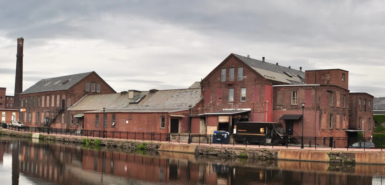 A row of red-brick mill buildings along the First Level Canal at Holyoke Heritage State Park on an overcast afternoon — the Wauregan paper mill on the right, a gable-and-shed-roofed run of older mill buildings stretching to the left, a tall white-painted brick smokestack at the far left, the buildings reflected in the dark green water of the canal in the foreground, a black wrought-iron rail along the canal walkway, and a UPS delivery truck visible on an access lane between the buildings.